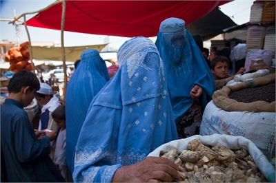 [ai] Two women wearing blue burqas in a market, with one woman reaching towards bags of goods. Children and other shoppers are visible in the background under colorful market awnings.